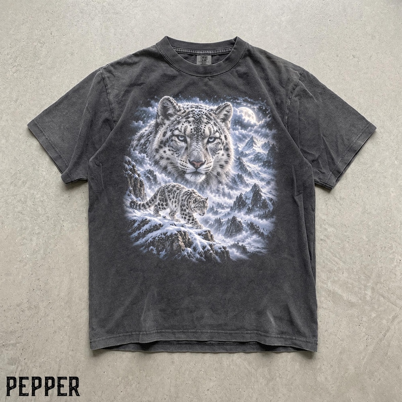 K&ouml;nnte beinhalten: Ein anthrazitfarbenes Kurzarm-T-Shirt mit einem Grafikdruck eines Schneeleopardengesichts und einem kleineren Schneeleoparden, der unter einem Vollmond &uuml;ber ein verschneites Gebirge l&auml;uft. Das Shirt hat einen Rundhalsausschnitt und eine verwaschene Optik.