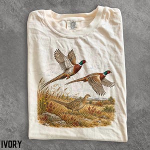 Puede incluir: Una camiseta de color marfil con un estampado gráfico de tres faisanes en un campo de hierba. Dos faisanes aparecen en vuelo con las alas extendidas, mientras que un faisán está de pie en el suelo. La camiseta tiene cuello redondo y una etiqueta Comfort Colors.