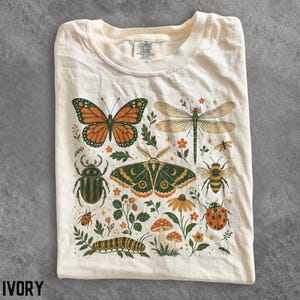 May include: An ivory t-shirt featuring a graphic print of various insects and plants. The design includes a monarch butterfly, a dragonfly, a moth, a beetle, a bee, a ladybug, a caterpillar, mushrooms, and small flowers in orange and green tones.