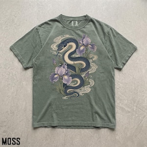 May include: A sage green short-sleeve t-shirt featuring a central graphic of a dark blue coiled snake surrounded by purple iris flowers and swirling cream-colored smoke patterns. The shirt has a crew neckline and a standard fit.