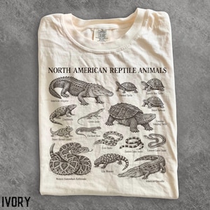 May include: A cream-coloured t-shirt featuring a dark brown graphic print titled NORTH AMERICAN REPTILE ANIMALS. The design displays detailed illustrations of various reptiles including an American alligator, turtles, snakes, and lizards with labels.