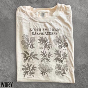 Puede incluir: Una camiseta color marfil con un estampado botánico marrón oscuro titulado North American Oaks & Acorns. El gráfico muestra nueve ilustraciones distintas de hojas de roble y bellotas con etiquetas como White Oak, Northern Red Oak y Bur Oak.