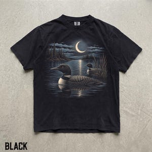 May include: A black short-sleeved t-shirt featuring a graphic print of two loons swimming on a lake at night. The scene includes a crescent moon in the sky, dark pine trees along the shoreline, and tall reeds emerging from the water's edge.