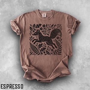 Puede incluir: Una camiseta de manga corta y cuello redondo en color marrón apagado. El centro de la prenda presenta un estampado gráfico negro de un zorro saltando a través de un denso arreglo de hojas de roble, bellotas, helechos y flores pequeñas. La etiqueta dice Comfort Colors.