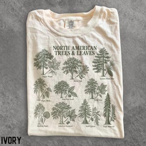 May include: An ivory t-shirt featuring a dark green graphic print titled North American Trees & Leaves. The design displays illustrations of various trees and their corresponding leaves, including Sugar Maple, White Oak, and Bald Cypress.
