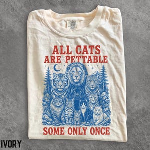 Può includere: Una t-shirt color avorio con un'illustrazione grafica blu di vari gatti selvatici e domestici, tra cui un leone, una tigre e un leopardo, circondati da alberi e una luna crescente. Il testo rosso recita ALL CATS ARE PETTABLE SOME ONLY ONCE.