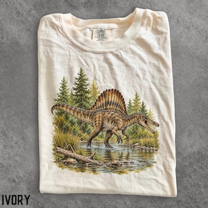 Op de afbeelding: Een ivoorkleurig t-shirt met een grafische print van een bruine en beige Spinosaurus die in ondiep water staat. De achtergrond toont een bos met groene dennenbomen en een boomstam. Het t-shirt heeft een Comfort Colors merklabel in de nek.