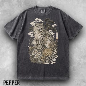 以下が含まれることがあります： チャコールグレーの半袖Tシャツ。岩の上に座るユキヒョウと、その周りに描かれた花、雲、松の木がグラフィックプリントされています。右側には縦書きの日本語の文字がデザインの一部として含まれています。