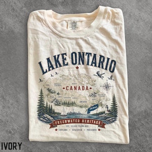 以下が含まれることがあります： アイボリー色のTシャツで、カナダの地図と「Lake Ontario」「Canada」「Freshwater Heritage」の文字が描かれています。デザインには、松の木、ヨット、灯台、鳥、コンパスローズのイラストが青と赤で施されています。
