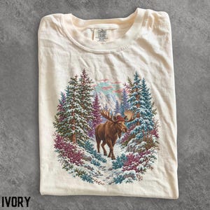 May include: An ivory t-shirt featuring a graphic print of a brown moose wearing a red beanie, standing in a snowy forest with evergreen trees and distant mountains. The shirt has a crew neckline and a Comfort Colors brand label at the collar.