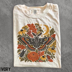 May include: An ivory t-shirt featuring a graphic print of a large moth centered among autumn-toned foliage, yellow flowers, a crescent moon, a beetle, and a ladybug. The shirt has a crew neckline and a Comfort Colors brand label at the inner collar.