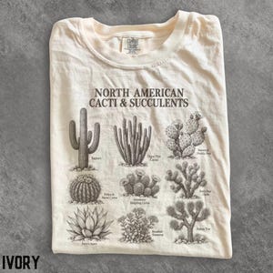 Puede incluir: Una camiseta color marfil con un estampado botánico marrón oscuro titulado North American Cacti & Succulents. El gráfico muestra ilustraciones de Saguaro, cactus órgano, nopal, cactus barril, cactus erizo, cholla, agave, sedum y árbol de Josué.