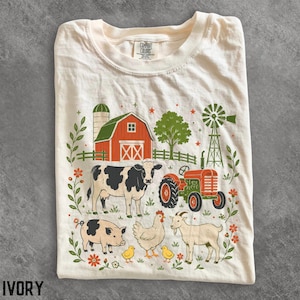 以下が含まれることがあります： アイボリー色のTシャツで、農場の風景のグラフィックプリントが施されています。赤い納屋、緑の風車、白黒の牛、豚、鶏、2羽のひよこ、ヤギが描かれており、周囲には緑の葉と小さなオレンジ色の花が配置されています。