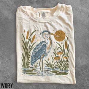 以下が含まれることがあります： アイボリー色のTシャツ。水辺に立つ青と灰色のサギ、ガマ、スイレンの葉、花が描かれたグラフィックプリントが特徴。質感のある金色の太陽と小さな星が鳥を囲んでいる。首元の内側のラベルにはComfort Colorsと記載されている。