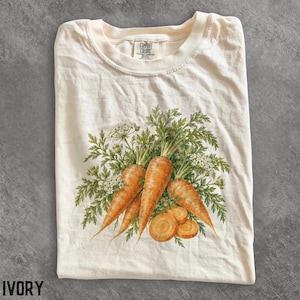 Könnte beinhalten: Ein elfenbeinfarbenes T-Shirt mit einer detaillierten botanischen Illustration von orangefarbenen Karotten mit grünem Blattwerk und kleinen weißen Blüten. Das Motiv ist mittig platziert und das Shirt hat ein Comfort Colors Nackenetikett.