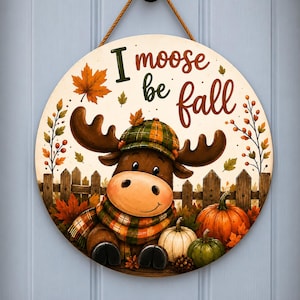 May include: A round wooden sign featuring a cartoon moose wearing a plaid hat and scarf, standing before a wooden fence with pumpkins and autumn leaves. The text "I moose be fall" is printed in dark green and brown script at the top of the sign.