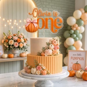 Può includere: Una torta a due piani decorata con piccole zucche e fiori, sormontata da un topper in acrilico con la scritta "Olivia is one" in lettere arancioni, che include un'illustrazione di una zucca con un fiocco rosa.