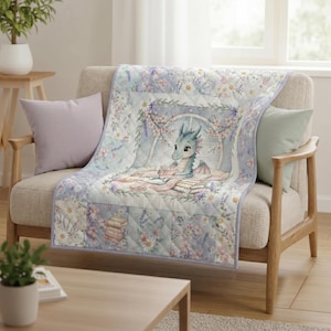 May include: A quilted throw blanket featuring a light blue dragon reading a book, surrounded by floral patterns, daisies, and lavender. The design includes a central illustration of the dragon in a window frame with stacks of books and tea cups.
