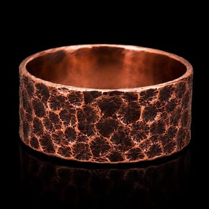 Op de afbeelding: Een brede ring gemaakt van koper, voorzien van een zwaar gehamerd, getextureerd oppervlak met een donkere, geoxideerde afwerking die de diepe inkepingen en verhoogde metalen ribbels over de gehele buitenkant van de ring benadrukt.