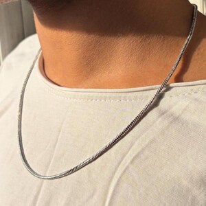 May include: A silver-toned snake chain necklace with a smooth, flexible, and rounded link design. The metallic chain rests against a plain white fabric surface, showcasing a consistent, tightly woven texture throughout the entire length of the piece.