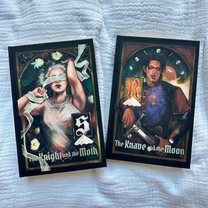 Op de afbeelding: Twee hardcover boeken met geïllustreerde covers. Het linkerboek, getiteld The Knight and the Moth, toont een geblinddoekte figuur met witte linten en motten. Het rechterboek, The Knave and the Moon, toont een figuur die een zwaard vasthoudt.