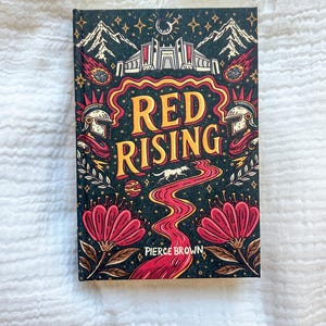 May include: A hardcover book titled Red Rising by Pierce Brown with a dark cover featuring gold text, a central red winding path, a white wolf, two helmets, stylized mountains, and large red flowers against a black background with stars.