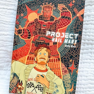 May include: A hardcover book titled Project Hail Mary by Andy Weir. The cover features a mosaic-style illustration of an astronaut in a red suit and a man in a cream-colored cardigan with a shirt that reads Horse Shoe Auto Club.