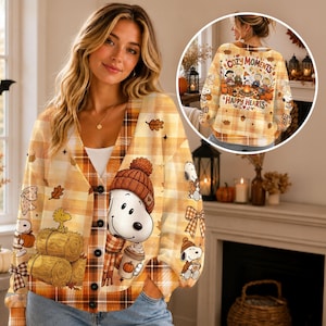 Peut inclure: Un cardigan boutonné à carreaux beige et orange orné de Snoopy portant un bonnet en tricot et une écharpe, tenant une boisson à la citrouille. Le motif inclut des bottes de foin, des feuilles d'automne et des nœuds. Un encart montre le dos avec le texte Cozy Moments Happy Hearts.