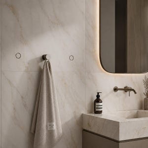 Op de afbeelding: Een moderne badkamer met een beige handdoek die aan een chromen haak aan een muur met marmerlook hangt. Een rechthoekige spiegel met een donker frame is zichtbaar, samen met een wastafel en een zeepdispenser. De handdoek heeft de tekst "HILA".
