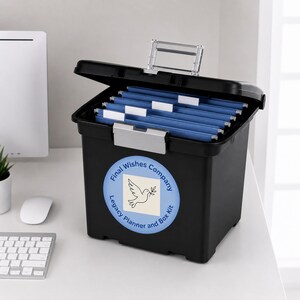 May include: Black file box with an open lid, containing blue hanging file folders. The box has a label that reads "Final Wishes Company Legacy Planner and Box Kit" with a dove graphic. The box is on a white desk.