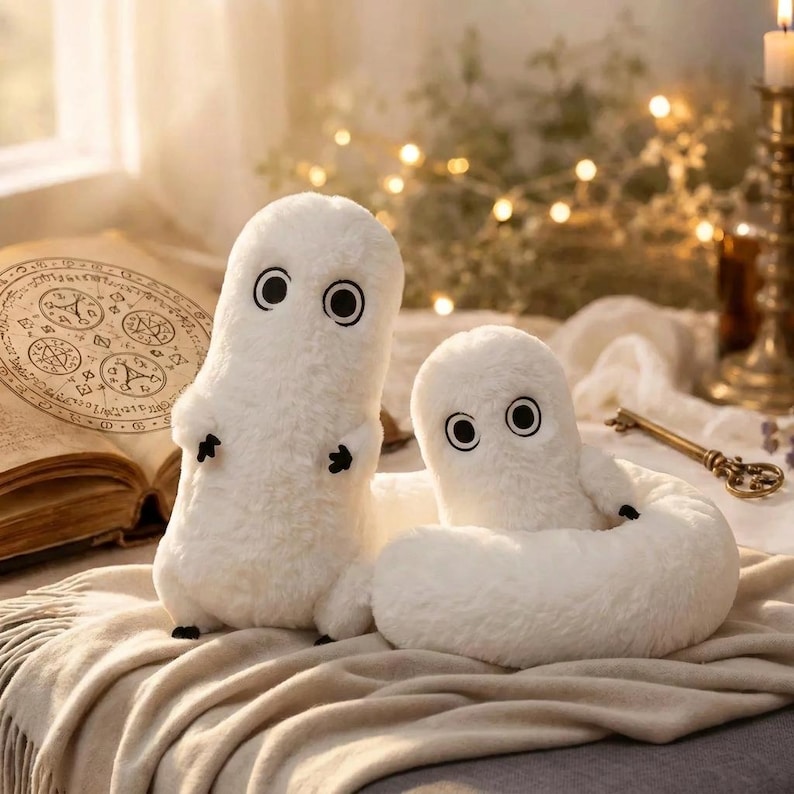 Puede incluir: Dos peluches blancos con ojos negros y peque&ntilde;os ap&eacute;ndices negros. Un juguete est&aacute; erguido, el otro est&aacute; enrollado. Est&aacute;n sobre una manta beige, con un libro y una vela en el fondo. Los juguetes tienen una textura suave y difusa.