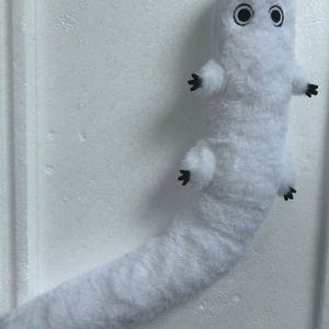 Puede incluir: Un peluche blanco con forma de gusano o oruga. Tiene una cabeza redondeada con dos ojos grandes con borde negro y peque&ntilde;os pies negros. El cuerpo es largo y esponjoso.
