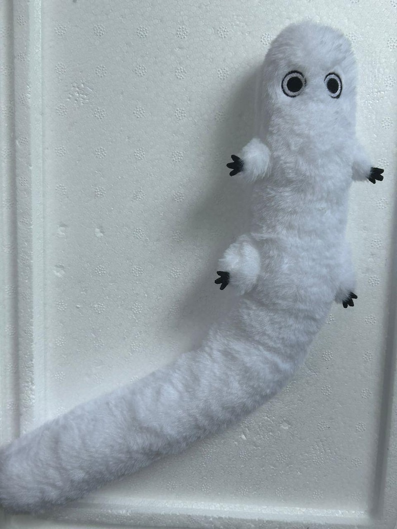 Puede incluir: Un peluche blanco con forma de gusano o oruga. Tiene una cabeza redondeada con dos ojos grandes con borde negro y peque&ntilde;os pies negros. El cuerpo es largo y esponjoso.