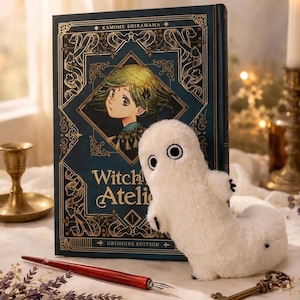Puede incluir: Un juguete de fantasma blanco de peluche con ojos negros y peque&ntilde;as garras negras est&aacute; junto a un libro de tapa dura. El libro tiene una cubierta azul oscuro con filigranas doradas y el t&iacute;tulo "Witch Atelier". Tambi&eacute;n se ven un bol&iacute;grafo rojo y una llave.