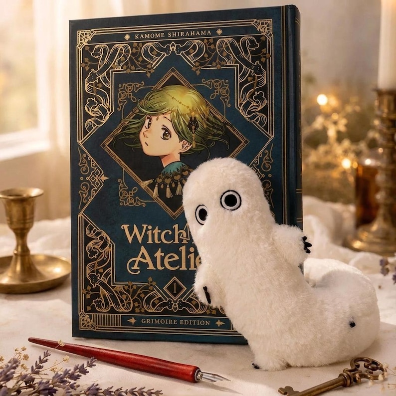 Puede incluir: Un juguete de fantasma blanco de peluche con ojos negros y peque&ntilde;as garras negras est&aacute; junto a un libro de tapa dura. El libro tiene una cubierta azul oscuro con filigranas doradas y el t&iacute;tulo "Witch Atelier". Tambi&eacute;n se ven un bol&iacute;grafo rojo y una llave.