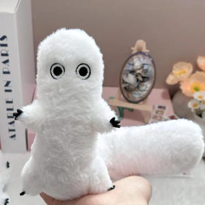 Puede incluir: Un peluche blanco con forma de peque&ntilde;a criatura esponjosa, con grandes ojos negros redondos y peque&ntilde;as manos y pies negros. El juguete tiene una larga cola cil&iacute;ndrica.