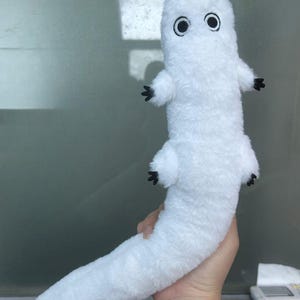 Puede incluir: Un peluche blanco con forma de gusano o oruga, con ojos negros y peque&ntilde;os pies negros. El juguete est&aacute; hecho de un material suave y esponjoso y tiene un cuerpo largo y curvado. El juguete est&aacute; sostenido por una mano.