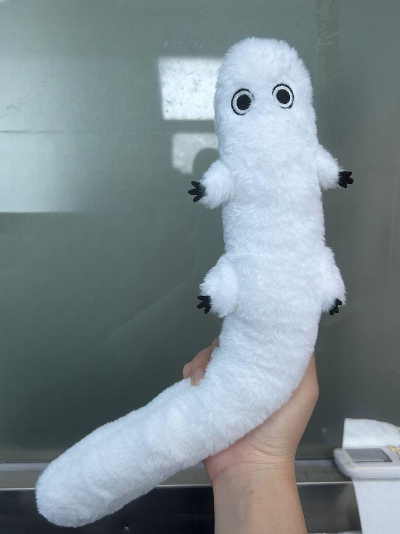 Puede incluir: Un peluche blanco con forma de gusano o oruga, con ojos negros y peque&ntilde;os pies negros. El juguete est&aacute; hecho de un material suave y esponjoso y tiene un cuerpo largo y curvado. El juguete est&aacute; sostenido por una mano.
