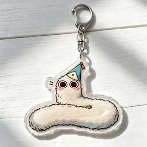 Puede incluir: Un llavero de acr&iacute;lico transparente con un gusano de dibujos animados que lleva un gorro de fiesta azul. El gusano es blanco con una textura esponjosa, ojos grandes y mejillas rosadas. El llavero tiene un cierre de metal plateado.