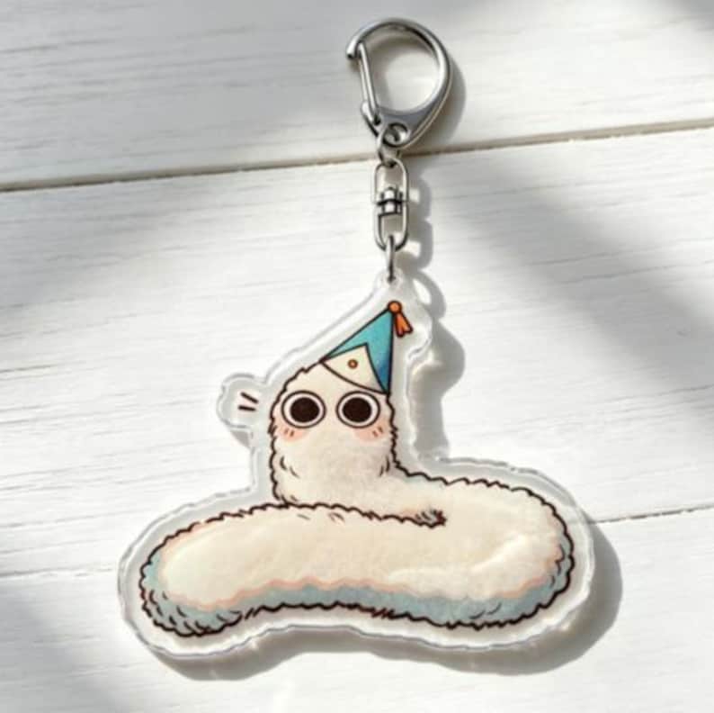 Puede incluir: Un llavero de acr&iacute;lico transparente con un gusano de dibujos animados que lleva un gorro de fiesta azul. El gusano es blanco con una textura esponjosa, ojos grandes y mejillas rosadas. El llavero tiene un cierre de metal plateado.