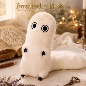 Könnte beinhalten: Ein weißes Plüschtier mit großen schwarzen Augen und kleinen schwarzen Füßen. Der Text "Brushbuddy Plush" steht darüber. Das Plüschtier liegt auf einem Tisch mit einem Buch und einem Schlüssel.
