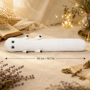 Puede incluir: Un peluche blanco con forma de lagarto, con ojos negros y peque&ntilde;as patas negras. El juguete mide aproximadamente 40 cm (15,7 pulgadas) de largo. El peluche est&aacute; sobre una superficie clara.