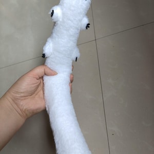 Puede incluir: Un peluche blanco con forma de gusano o oruga, con ojos negros y peque&ntilde;os ap&eacute;ndices negros. El juguete se sostiene en una mano y parece suave y esponjoso.