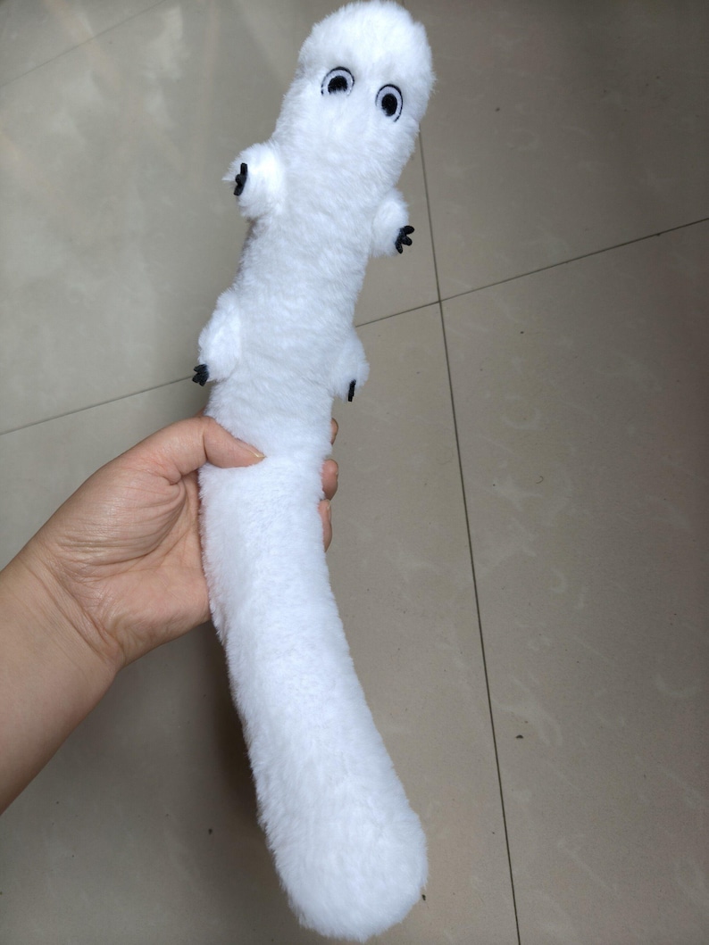Puede incluir: Un peluche blanco con forma de gusano o oruga, con ojos negros y peque&ntilde;os ap&eacute;ndices negros. El juguete se sostiene en una mano y parece suave y esponjoso.