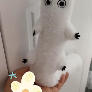 Puede incluir: Un peluche blanco con forma de criatura, con grandes ojos negros y peque&ntilde;as manos y pies negros. El juguete se sostiene en una mano, con una pared blanca de fondo.
