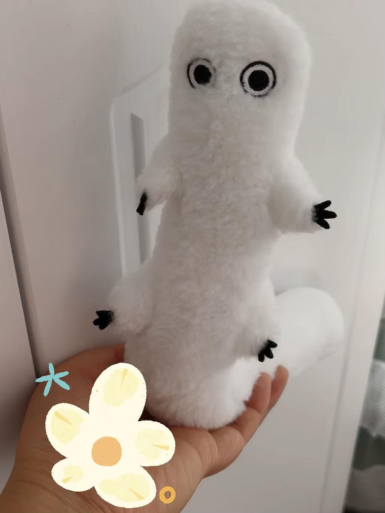 Puede incluir: Un peluche blanco con forma de criatura, con grandes ojos negros y peque&ntilde;as manos y pies negros. El juguete se sostiene en una mano, con una pared blanca de fondo.