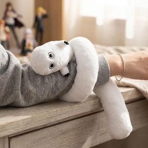 Puede incluir: Un juguete de peluche blanco con forma de gusano o serpiente, con ojos negros y peque&ntilde;os detalles negros. El juguete est&aacute; envuelto alrededor de una manga gris. El juguete est&aacute; sobre una superficie de madera.
