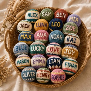 May include: A collection of colorful, crocheted balls with names embroidered on them, displayed in a woven basket. Each ball features a different name in a contrasting color, such as "ALEX", "SAM", and "BEN". The balls are in various colors, including blue, green, and yellow.