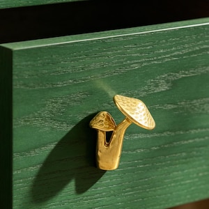 May include: A gold-toned metal cabinet knob shaped like two mushrooms with textured caps, attached to a dark green wooden drawer front with a visible wood grain pattern and a smooth, matte finish on the drawer surface.