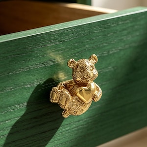 Op de afbeelding: Een goudkleurige metalen ladeknop in de vorm van een teddybeer die een glad hart vasthoudt. De knop is bevestigd aan een donkergroene houten ladefront met een zichtbare houtnerftextuur, voorzien van een horizontaal ontwerp en een open lade.